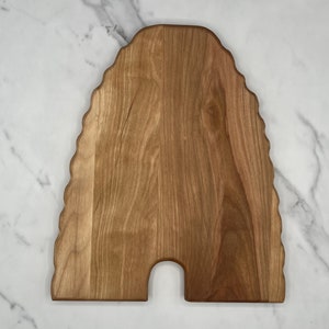 May include: A wooden cutting board in the shape of a beehive. The board is made of light brown wood with a natural grain pattern. It has a scalloped edge and a small, arched cutout at the bottom, resembling a hive entrance.