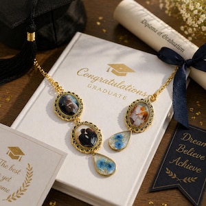May include: A gold-toned necklace with oval pendants holding small photos, and teardrop-shaped blue floral resin drops. The necklace rests on a white book with "Congratulations Graduate" text, next to a graduation cap and diploma.