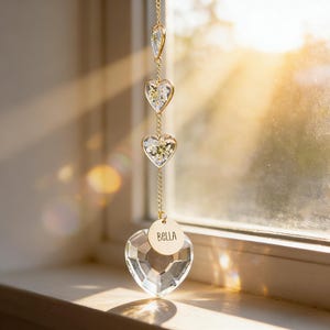 May include: A decorative suncatcher with a gold chain and clear crystal heart-shaped pendants. The suncatcher has a small gold tag that reads "Bella". The pendants are filled with small white flowers.