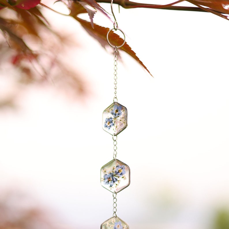 May include: A hanging ornament featuring three clear hexagonal resin charms connected by a silver-toned metal chain. Each charm contains pressed blue and white flowers embedded within the transparent material, suspended from a silver hook.