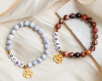 May include: Two beaded bracelets with gold paw charm accents. One bracelet is white with black marbling and spells "DUDU". The other bracelet is brown, pink, and black with gold beads and spells "JOJO".