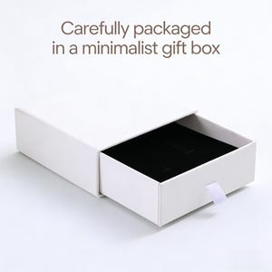 May include: A white rectangular sliding gift box with a black velvet interior insert and a small white pull tab. The text "Carefully packaged in a minimalist gift box" is printed in brown above the box on a plain white background.