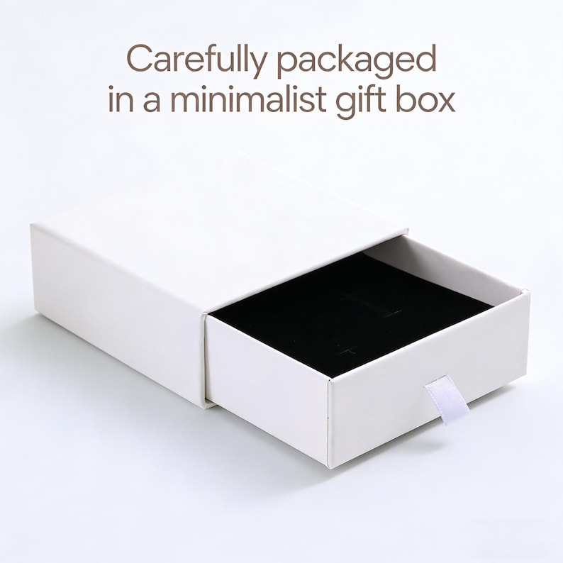 May include: A white rectangular sliding gift box with a black velvet interior insert and a small white pull tab. The text "Carefully packaged in a minimalist gift box" is printed in brown above the box on a plain white background.