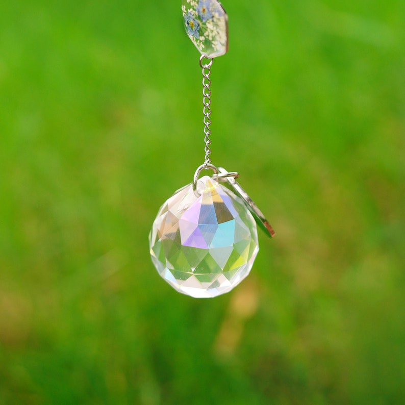 May include: A faceted clear crystal sphere with an iridescent finish suspended by a silver-toned metal chain. Attached to the top of the chain is a small hexagonal charm containing pressed blue flowers and white foliage encased in clear resin.