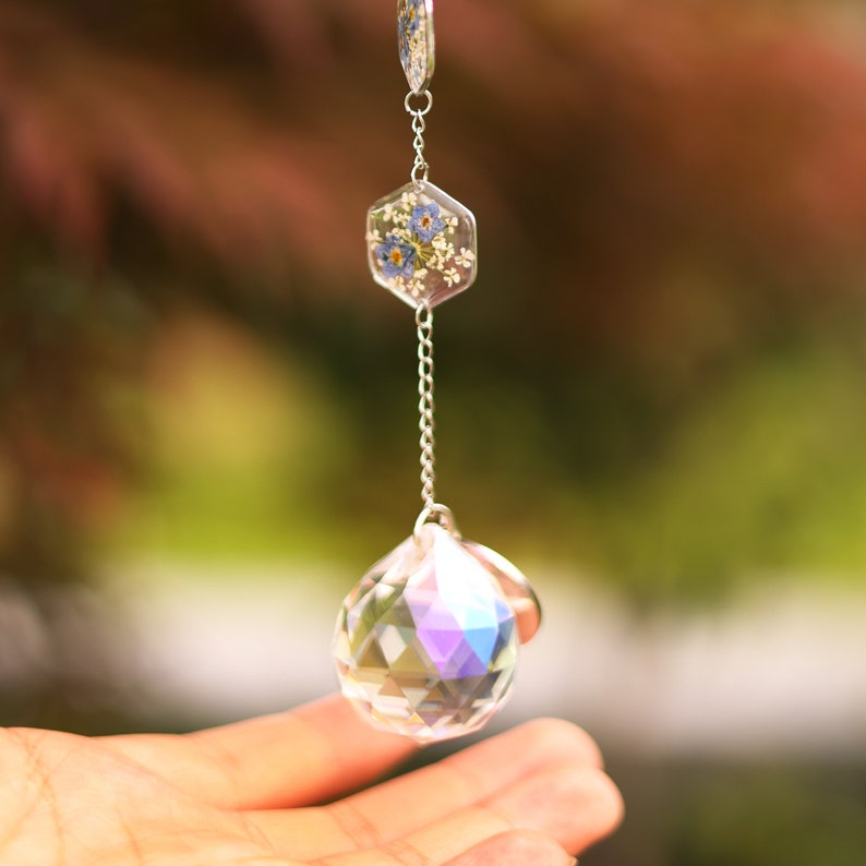 May include: A hanging suncatcher ornament featuring a faceted clear crystal sphere at the bottom, connected by a silver-toned metal chain to a small hexagonal resin charm containing embedded blue and white pressed flowers.