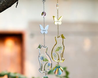 May include: Two hanging suncatchers featuring cat-shaped resin pendants filled with pressed flowers and greenery. Each chain includes a sun charm, a butterfly charm with the text LULU, and a faceted iridescent crystal drop at the bottom.