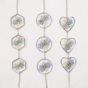 May include: Three vertical silver-toned chains featuring pressed blue forget-me-not flowers and white baby's breath encased in clear resin. The pendants are shaped as hexagons, scalloped circles, and hearts, connected by delicate metal links.