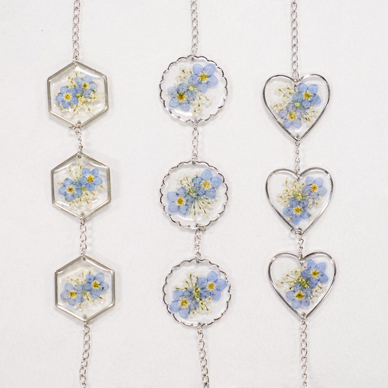 May include: Three vertical silver-toned chains featuring pressed blue forget-me-not flowers and white baby's breath encased in clear resin. The pendants are shaped as hexagons, scalloped circles, and hearts, connected by delicate metal links.