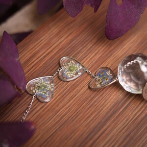May include: A silver-toned chain necklace featuring three heart-shaped pendants, each containing pressed blue and white flowers encased in clear resin, connected to a faceted clear crystal bead at the end of the chain structure.