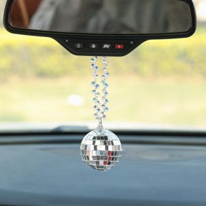 May include: A silver disco ball car charm hanging from a clear beaded chain. The charm is hanging from the rearview mirror of a car.