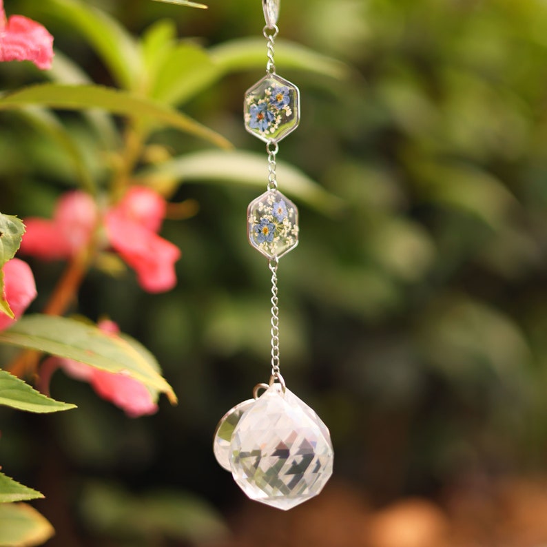 May include: A hanging suncatcher featuring two clear hexagonal resin charms containing pressed blue forget-me-not flowers and white foliage, connected by a silver-toned metal chain to a faceted clear glass crystal sphere at the bottom.