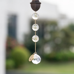 Puede incluir: Un atrapasol de cristal con una cadena dorada y tres cuentas redondas de cristal con flores prensadas en su interior. El atrapasol tiene una pequeña etiqueta dorada con la palabra "LUCK" en ella.
