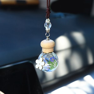 Może przedstawiać: Przezroczysta szklana butelka z drewnianym korkiem i małym białym kwiatem zawieszonym na brązowym sznurku. Butelka jest wypełniona suszonymi niebieskimi kwiatami i zielenią.