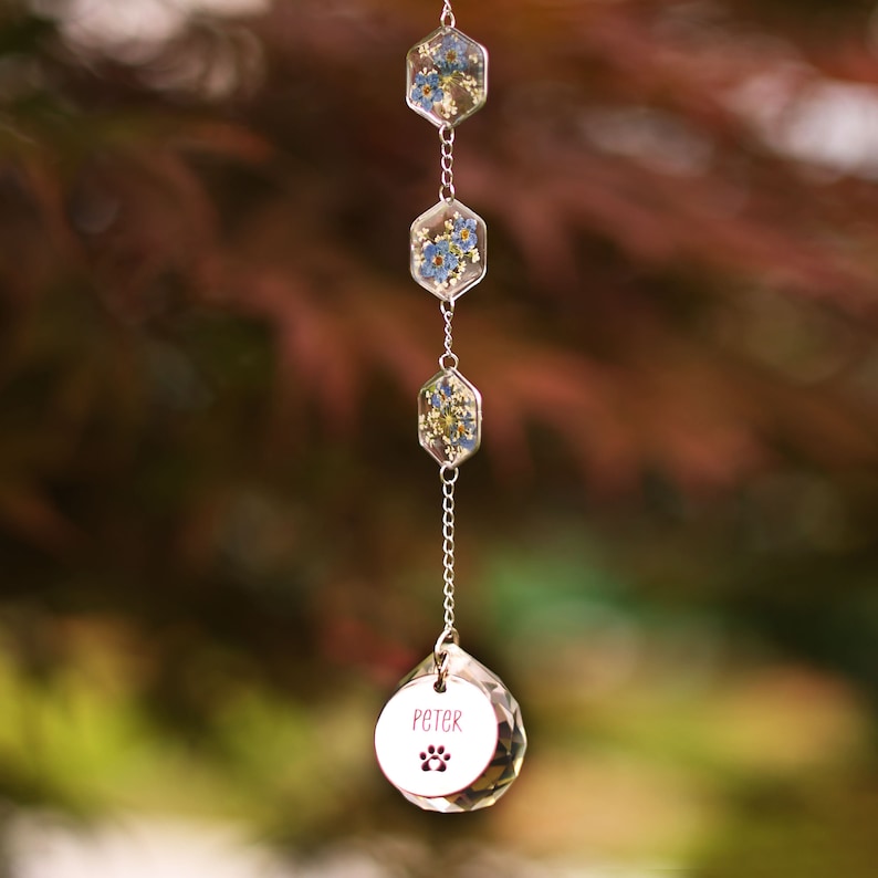 May include: A hanging suncatcher ornament featuring three vertical hexagonal resin charms containing pressed blue and yellow flowers, connected by silver chains to a clear faceted crystal sphere engraved with the name Peter and a paw print.