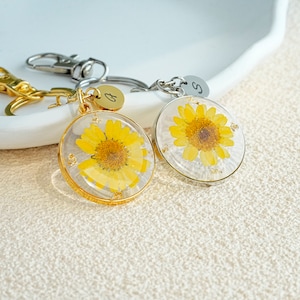 May include: Two gold and silver keychains with a small round charm featuring a pressed yellow flower. The charms are attached to a keyring with a letter initial.