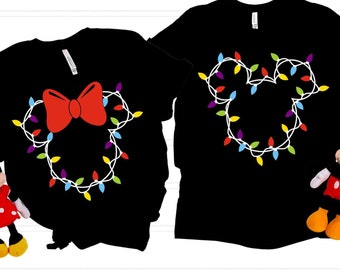 Pode incluir: Duas camisetas pretas com um design de luzes de Natal em forma de cabeça do Mickey Mouse. Uma camiseta tem um laço vermelho nas luzes, a outra não.