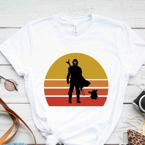 May include: White t-shirt with a retro sunset graphic featuring a silhouette of a Mandalorian and Grogu.