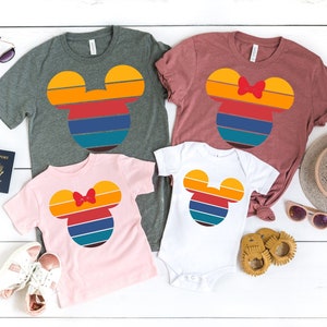 Peut inclure: Un ensemble de quatre t-shirts avec un motif de tête de Mickey Mouse de style rétro. Les t-shirts sont de différentes couleurs, dont le gris, le rose, le blanc et le marron. Le motif présente un arc-en-ciel de couleurs avec un contour jaune et un nœud rouge sur le t-shirt rose.