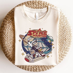 May include: A white t-shirt with a vintage-style graphic of Mickey Mouse and friends riding Space Mountain. The graphic features the words "Space Mountain" in red and white.