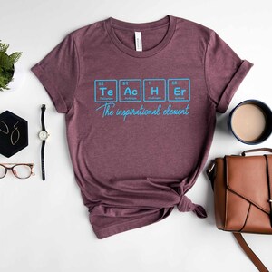 May include: A maroon t-shirt with a graphic of the periodic table elements spelling out "Teacher" and the text "The inspirational element".