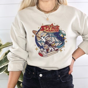 May include: A beige sweatshirt with a graphic print of a cartoon space mountain ride with the text "Space Mountain" in red and white. The graphic features three cartoon characters riding the ride.