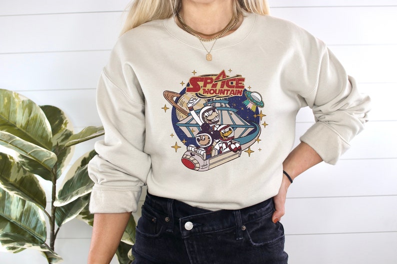 May include: A beige sweatshirt with a graphic print of a cartoon space mountain ride with the text "Space Mountain" in red and white. The graphic features three cartoon characters riding the ride.
