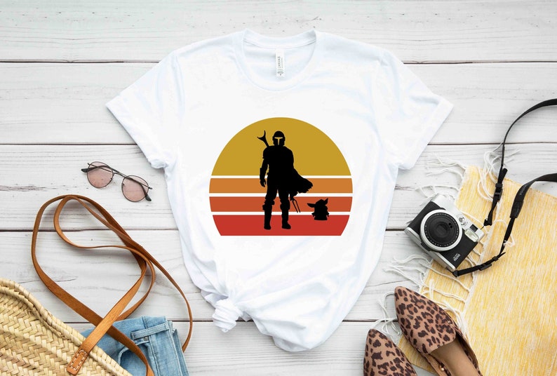 May include: White t-shirt with a retro sunset graphic featuring a silhouette of a Mandalorian and Grogu.