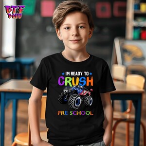 May include: Black t-shirt with the text "I'M READY TO CRUSH PRESCHOOL" in colorful letters. A monster truck graphic with red, white, and blue accents is featured below the text. The shirt is designed for children.