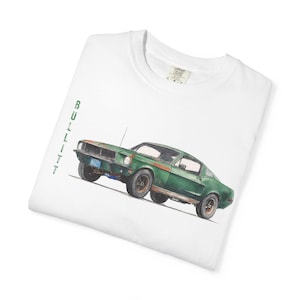 Puede incluir: Camiseta blanca con una ilustración de un coche verde vintage con detalles en color óxido. La palabra "BULITT" está impresa verticalmente en verde en el lado izquierdo. Cuello redondo y etiqueta.