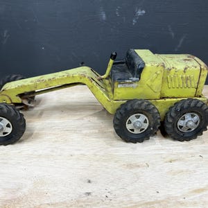 May include: A vintage yellow metal toy road grader featuring a black seat, a long front frame, and six black rubber tires with silver-colored hubs. The construction vehicle toy shows signs of wear and paint chipping on its yellow body.