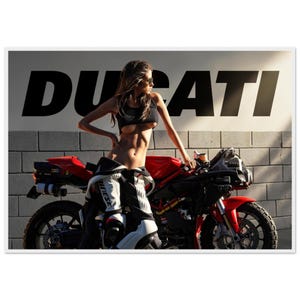 May include: A red Ducati motorcycle parked in front of a wall with the word DUCATI in large black letters. A person stands beside the motorcycle wearing a black crop top and white and black motorcycle racing trousers with Dainese branding.