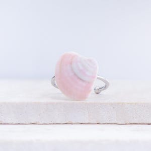Op de afbeelding: Een schelpenring met een lichtroze en witte schelp. De schelp is gemonteerd op een zilverkleurige verstelbare ringband. De ring wordt gepresenteerd op een wit, getextureerd oppervlak.