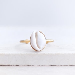 May include: A gold ring with a white seashell as its central feature. The oval-shaped shell is set in a gold bezel. The ring's band is thin and gold, offering a minimalist, coastal design. The ring is suitable for everyday wear.