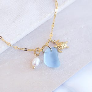 May include: A gold-coloured necklace with a delicate chain. The necklace features a light blue sea glass pendant, a small gold turtle charm, and a white pearl. The necklace is displayed on a white surface.