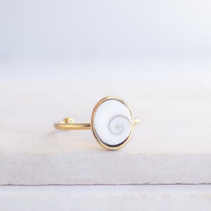 Pode incluir: Um anel dourado com um design oval branco em forma de espiral. O anel tem uma banda fina e um estilo simples e elegante. O design em espiral é centrado em uma moldura dourada.