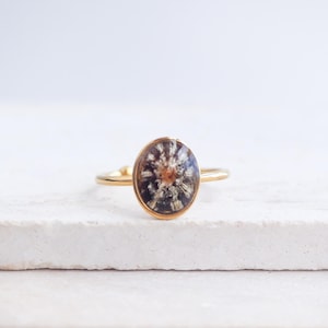 Peut inclure: Une bague dorée avec une pierre ovale. La pierre présente un motif de fleur séchée aux tons marron, blanc et noir. La bague est posée sur une surface blanche.