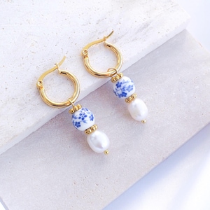 Peut inclure: Boucles d'oreilles créoles dorées avec perles en céramique florales bleues et blanches et gouttes de perles blanches. Les boucles d'oreilles sont présentées sur une surface blanche, mettant en valeur leur design élégant. Un accessoire de mode raffiné.