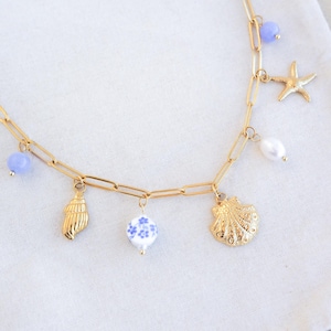 Può includere: Collana dorata con vari ciondoli, tra cui una stella marina, una conchiglia e una perla. La collana presenta anche perline blu e bianche. La catena ha un design a maglie allungate, creando un'estetica delicata e a tema mare.