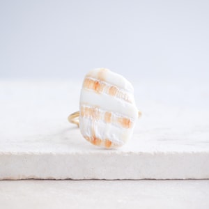 Puede incluir: Un anillo único con una concha marina blanca grande, de forma irregular, con rayas naranja-marrón. La concha está montada sobre una fina banda dorada. El anillo se muestra sobre un fondo neutro.