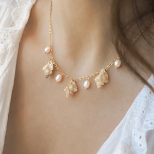Può includere: Una collana dorata con piccole conchiglie e perle bianche. La collana ha una catena delicata e si indossa intorno al collo. Le conchiglie sono di colore beige chiaro e le perle sono rotonde e lucenti. La collana è un gioiello.