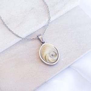 Può includere: Una collana argentata con un ciondolo ovale con un design a conchiglia naturale. Il ciondolo, incorniciato in argento, pende da una catena sottile. La conchiglia presenta un motivo a spirale nei toni crema e beige.