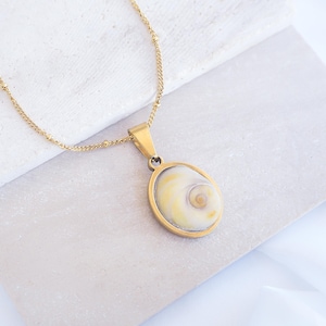 May include: A gold-colored necklace with a pendant featuring a seashell design. The pendant is oval-shaped with a gold frame and a cream and yellow seashell center. The chain is delicate and gold, resting on a light-colored surface.