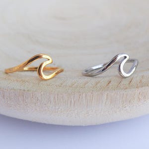 Peut inclure: Deux bagues en forme de vague, l'une dorée et l'autre argentée, sont présentées sur une surface en bois clair. Les bagues ont un design minimaliste, ressemblant à des vagues océaniques. La bague dorée est à gauche et la bague argentée à droite.