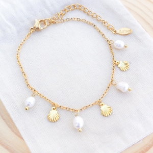 Pode incluir: Pulseira de corrente dourada com pingentes pendurados. A pulseira apresenta pequenos pingentes de conchas douradas e contas de pérolas brancas. A pulseira é exibida em um tecido branco, com um fundo de madeira.