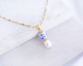 Könnte beinhalten: Eine goldene Halskette mit einem Anhänger, der aus einer kleinen, runden, weißen Keramikperle mit blauem Blumenmuster besteht, akzentuiert durch zwei goldfarbene Zwischenstücke und eine weiße, tropfenförmige Süßwasserperle am unteren Ende.