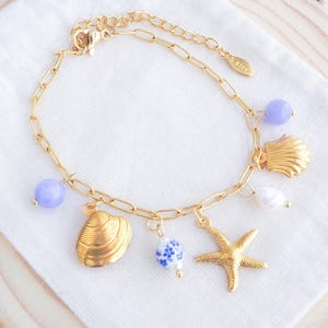 Op de afbeelding: Goudkleurige armband met een schakelkettingontwerp, versierd met schelp-, zeester- en kralenbedels. De armband bevat een lichtpaarse kraal en een witte parel. De bedels zijn goud en blauw.