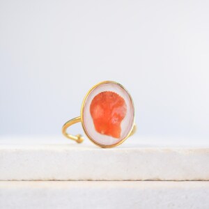 Pode incluir: Um anel dourado com uma moldura oval de resina transparente contendo um elemento orgânico laranja. O anel tem uma banda ajustável e uma fina borda dourada ao redor da moldura. O anel é exibido em uma superfície branca. A moldura mede aproximadamente 2 cm.