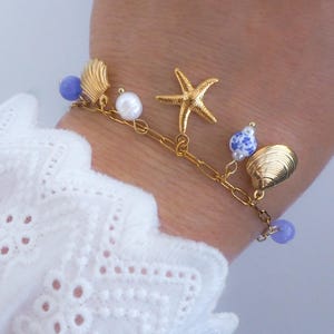 Pu&ograve; includere: Bracciale con ciondoli dorato con una stella marina, conchiglie e perline blu e bianche. Il bracciale &egrave; attaccato a una catena d'oro. Il bracciale &egrave; indossato su un polso, con una manica di pizzo bianco sullo sfondo.