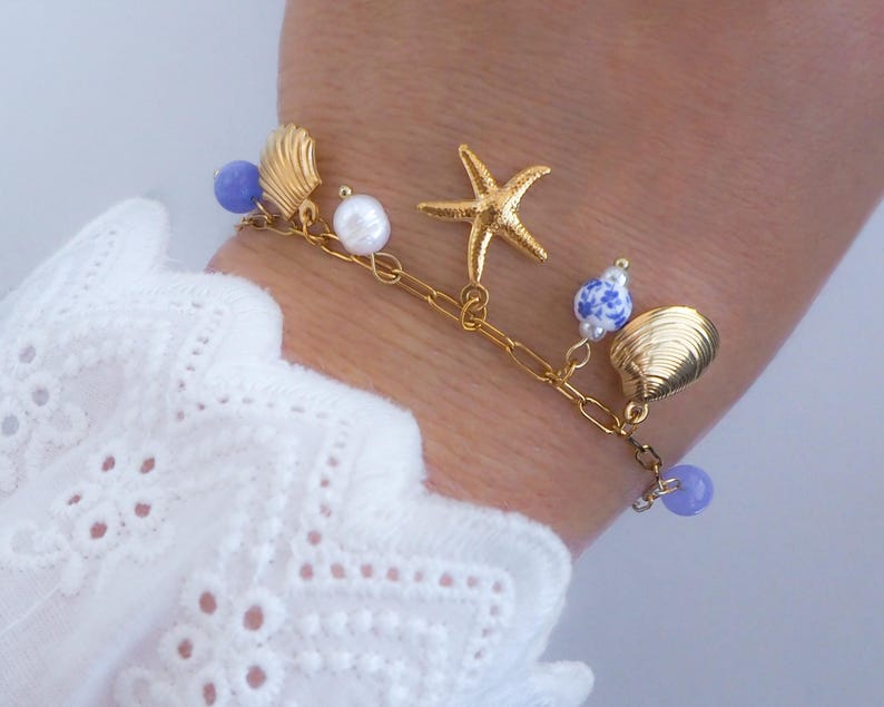 Pu&ograve; includere: Bracciale con ciondoli dorato con una stella marina, conchiglie e perline blu e bianche. Il bracciale &egrave; attaccato a una catena d'oro. Il bracciale &egrave; indossato su un polso, con una manica di pizzo bianco sullo sfondo.