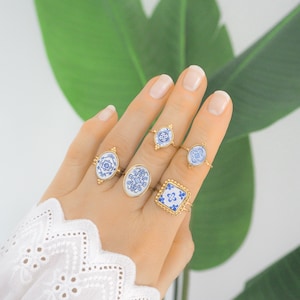Può includere: Un set di cinque anelli color oro con inserti in stile ceramica a motivi blu e bianchi. Gli anelli presentano design ovali, quadrati e a forma di diamante con intricati motivi floreali e geometrici su sottili fasce metalliche.